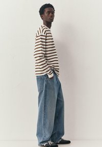 Ung mand, der står sidelæns iført en hvid og brun stribet langærmet skjorte, brede blå jeans og sorte sneakers med hvide snørebånd.