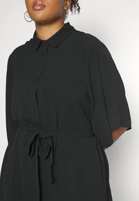 Femme portant une robe noire à manches courtes boutonnée, ceinture nouée à la taille et grandes boucles d'oreilles en anneau, cadrée aux épaules et au menton.