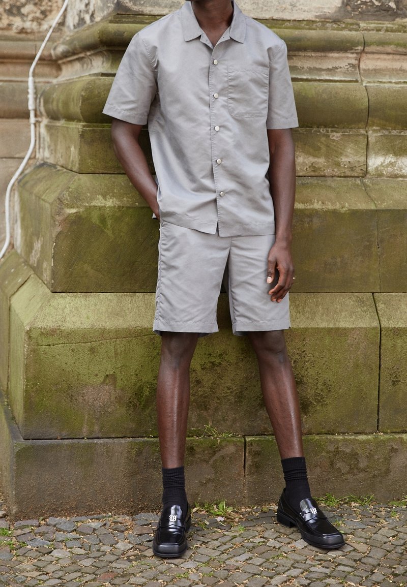 Grå kortärmad skjorta med knappar och matchande shorts, tillverkade av ett slätt tyg. Bärs med svarta loafers och strumpor, stående mot sten.