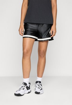 Person iført sorte træningsshorts med hvid kant, hvide crew-sokker og sort-hvide sneakers, der står mod en ensfarvet baggrund.