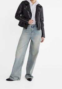 Schwarze Leder-Motorradjacke über einem grauen Kapuzenpullover, kombiniert mit weit geschnittenen, hellblauen Jeans und schwarzen Schuhen mit spitz zulaufender Spitze.