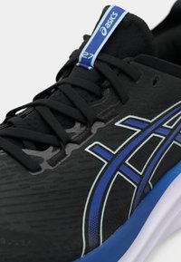 Chaussure de sport noire avec une surface texturée, arborant le logo Asics bleu et blanc, un système de laçage complexe et un accent bleu sur la semelle.