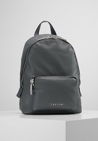 Sac à dos en cuir gris avec un dessus arrondi, poche zippée à l'avant et quincaillerie argentée. Doté de bretelles réglables et d'un logo en bas.
