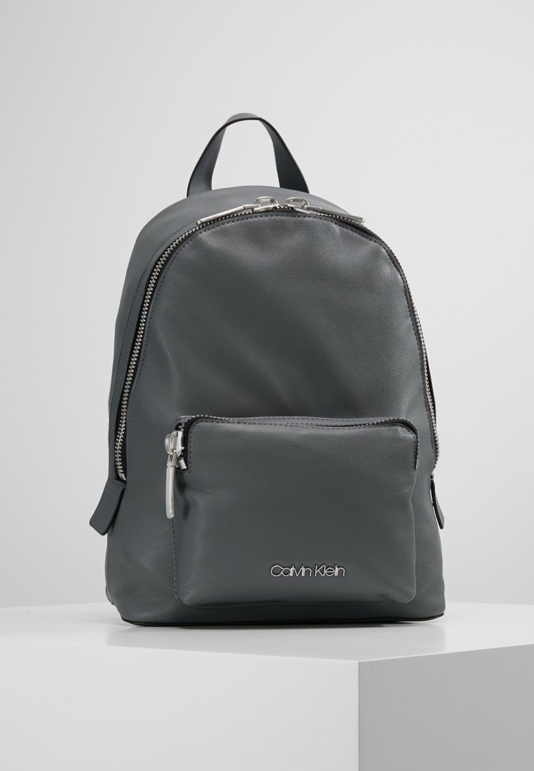 Sac à dos en cuir gris avec un dessus arrondi, poche zippée à l'avant et quincaillerie argentée. Doté de bretelles réglables et d'un logo en bas.