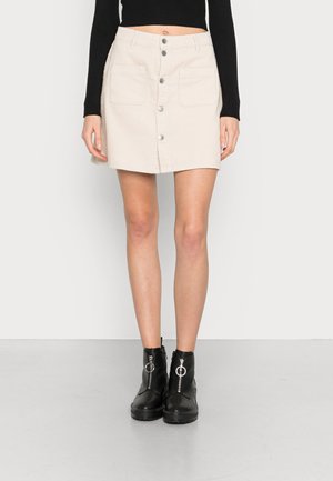 Jupe en velours côtelé beige clair avec fermeture à boutons sur le devant, deux grandes poches et un ourlet droit. Associée à des bottines noires.