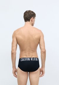 Čierne plavky s bielym logom "CALVIN KLEIN" na páse, vyrobené z hladkého materiálu, priliehavý strih a nízky rez.