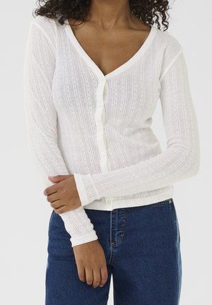 Frau trägt eine weiße, langärmelige, geknöpfte Strickjacke mit vertikalen Lochmustern und dunkelblaue Jeans aus Denim, die Arme vor der Taille verschränkt.