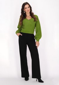 Grüner glitzernder Wickelpullover mit langen Puffärmeln, kombiniert mit hoch taillierten schwarzen Schlaghose und schwarzen High Heels.