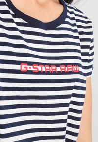 Camiseta a rayas en azul marino y blanco con cuello redondo. Presenta un logo rojo de "G-STAR RAW" en el frente. Mangas cortas y textura de algodón suave.