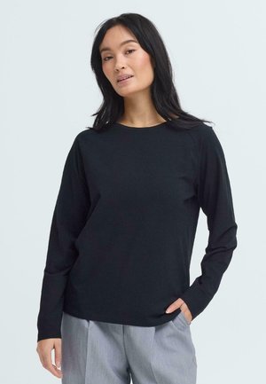 Vrouw met lang zwart haar, gekleed in een zwart shirt met lange mouwen en lichtgrijze broek, staand met één hand in de zak tegen een lichte achtergrond.