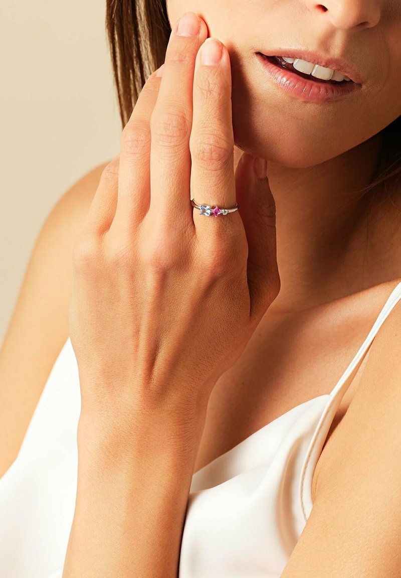 Silberring mit drei farbigen Steinen – zwei blau und einen pink – in einem zarten Band, getragen an einer Hand vor einem neutralen Hintergrund.