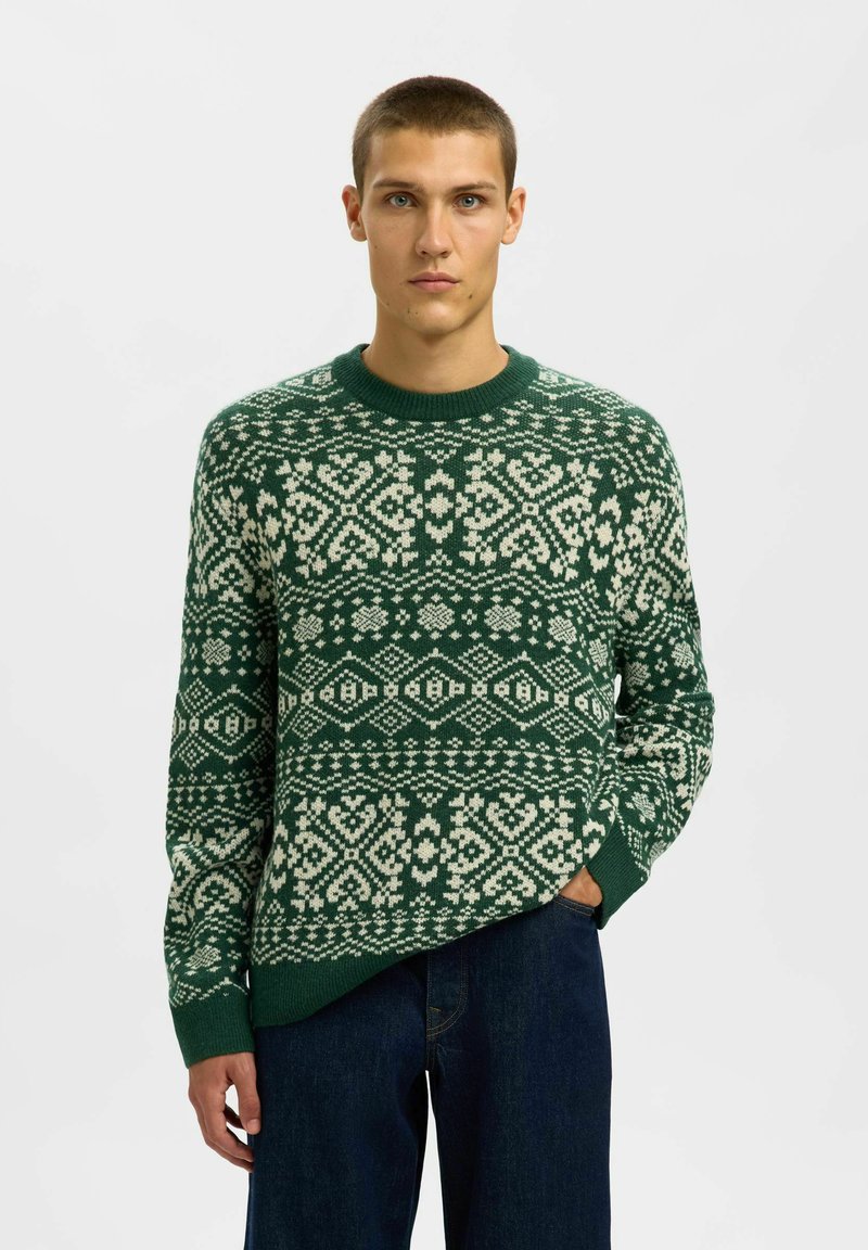 Maglione verde a maglia con un motivo geometrico bianco, scollatura tonda e polsini a costine, abbinato a jeans scuri in denim.