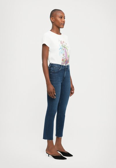 Witte graphic t-shirt met kleurrijke opdruk, gecombineerd met donkerblauwe enkelhoge spijkerbroek met een rafelige zoom. Zwarte sleehakken maken de look compleet.