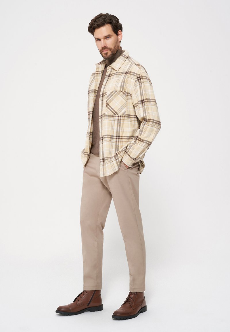 Beżowa koszula w kratę z brązowymi akcentami, brązowy golf, beżowe spodnie i brązowe skórzane botki. Koszula ma kieszeń na piersi.