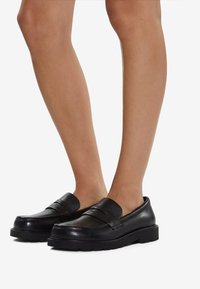 Svarta läderloafers med ett brett rem över framsidan, texturerad yta och tjock svart gummisula. Inga synliga dekorationer.