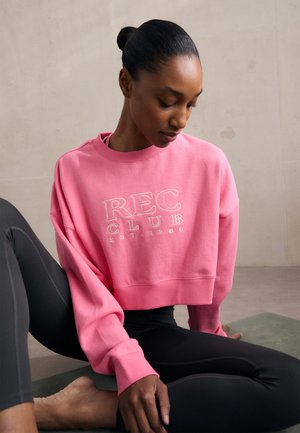 Sweatshirt rose court avec le logo "REC CLUB EST. 1980" en blanc. Équipé de manches larges et d'un col côtelé. Associé à des leggings noirs.