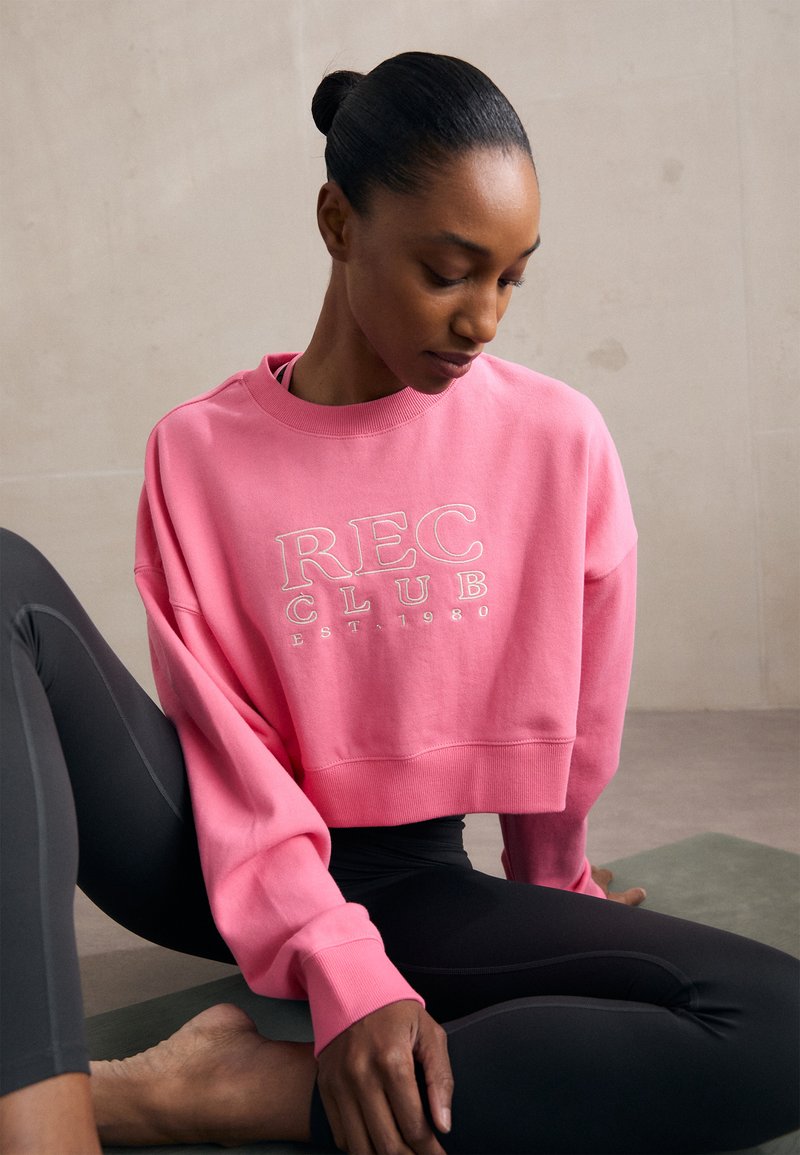 Rózsaszín, rövidített pulóver "REC CLUB EST. 1980" fehér logóval. Széles ujjakkal és bordázott nyakkal rendelkezik. Fekete leggings-szel párosítva.