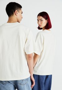 Krémové, oversized bavlněné trička nosí dva modely, s krátkými rukávy a kulatým výstřihem, doplněné o světle modré džíny.