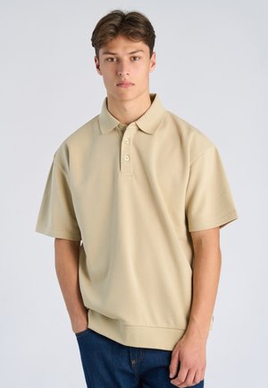 Ung man med ljust brunt hår, klädd i en beige pikétröja och blå jeans, står med ena handen i fickan mot en enfärgad bakgrund.