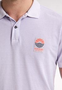 Světle fialová polo košile vyrobená z texturované látky, s límečkem a našitou nášivkou s designem slunce a vln v oranžové a námořnické modři.