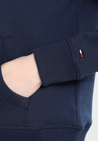 Navy blue sweatshirt with ribbed cuffs, a front pocket, and a small red, white, and blue logo accent on the sleeve. Soft cotton material.
