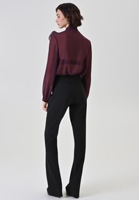 Vrouw staand opzij, gekleed in een doorschijnende donkerpaarse blouse en hooggetailleerde zwarte broek tegen een effen lichte achtergrond.