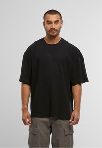T-shirt noir oversize à manches courtes avec une texture côtelée et un logo minimaliste sur le devant, associé à un pantalon cargo gris.