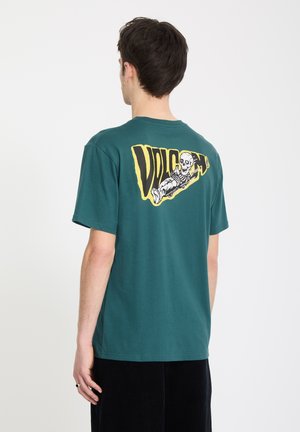 Man draagt een teal T-shirt met gele en zwarte afbeelding van een liggend skelet en grote tekst op de achterkant, kijkt weg van de camera.