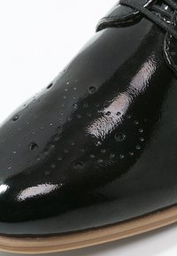 Chaussure en cuir verni noir avec détails perforés, bout arrondi et semelle extérieure beige. Présente une surface lisse et des lacets.
