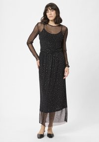 Melnā maxi kleita ar pieguļošu augšu, caurspīdīgām garām piedurknēm un izkaisītām sudraba punktiņām. Garums līdz potītēm ar caurspīdīgu slāni.