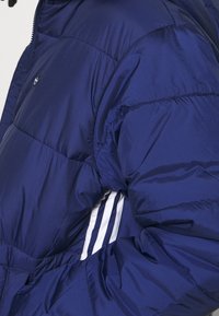 Marinblå puffjacka med blank yta, försedd med vita tre-stripes detaljer på ärmarna och en liten logotyp på bröstet.