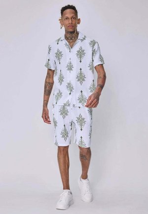 Homme tatoué portant un ensemble blanc assorti composé d'une chemise à manches courtes et d'un short avec des motifs géométriques verts et des baskets blanches, marchant vers l'avant.