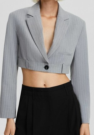 Woman wearing a cropped grey pinstripe blazer with one black button and high-waisted black pants, showing midriff and arms.