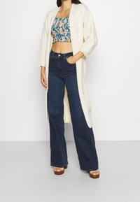 Krémový dlouhý kardigan, květinový zkrácený top v teplých a pastelových barvách, tmavé slim džíny a béžové platformové sandály. Jemné materi ály a uvolněný střih.