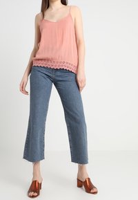 Top rosa senza maniche con orlo in pizzo, abbinato a jeans blu cropped e sabot con tacco in rame lucido. Texture morbida e vestibilità rilassata.