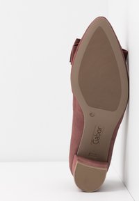 Chaussure en daim rose avec un bout pointu et un petit talon. La semelle est en caoutchouc beige avec une surface texturée et comporte des détails de logo de la marque.