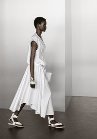 White sleeveless top and flowing white skirt, with structured fabric and asymmetrical hem. White open-toe sandals and a textured bag.