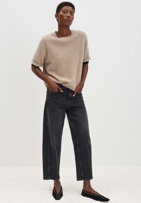 Persona che indossa un maglione beige a maniche corte con polsini arrotolati, jeans neri a gamba larga e ballerine nere con borchie, in piedi con le mani in tasca.