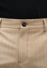 Μπεζ ριγέ παντελόνι με μαύρο κουμπί και θηλιές ζώνης, φορεμένο με μαύρο τοπ.