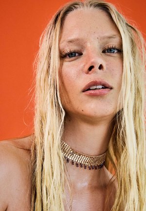 Zbliżenie na młodą kobietę z długimi blond włosami, piegami, lekkim makijażem i naszyjnikiem z koralików na pomarańczowym tle.