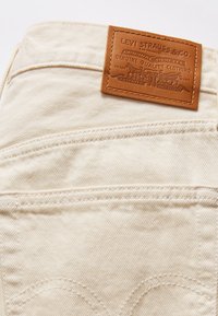Jean en denim crème avec une patch en cuir portant un logo gaufré. Détail de couture distinctif sur la poche arrière. Texture douce.