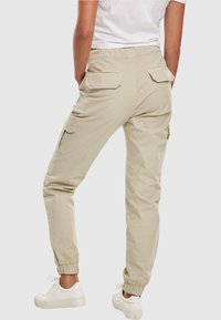 Osoba v béžových cargo kalhotách s elastickými manžetami a kapsami na klopy, v kombinaci s bílými teniskami a bílým tričkem, stojící na jednobarevném pozadí.