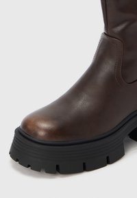 Rjavi usnjeni gležnjar z zaobljenim prstom in debelim črnim gumijastim podplatom z globokimi utori za oprijem. Gladka tekstura in minimalni detajli.