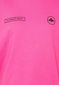 Rosa bomullströja med svart text som står "IKKE VANLIGE MENNESKER" och en cirkulär logga på höger sida. Mjuk textur, avslappnad design.