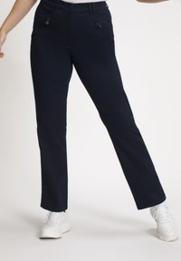 Personne portant un pantalon noir à jambes droites avec des poches avant zippées et des baskets blanches, debout les jambes écartées.