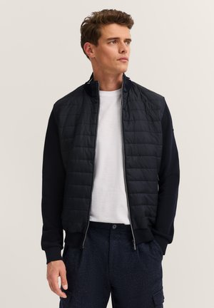 Junger Mann in einer schwarzen gesteppten Jacke über einem weißen T-Shirt und dunklen Hosen, steht mit den Händen in den Taschen und schaut nach rechts.