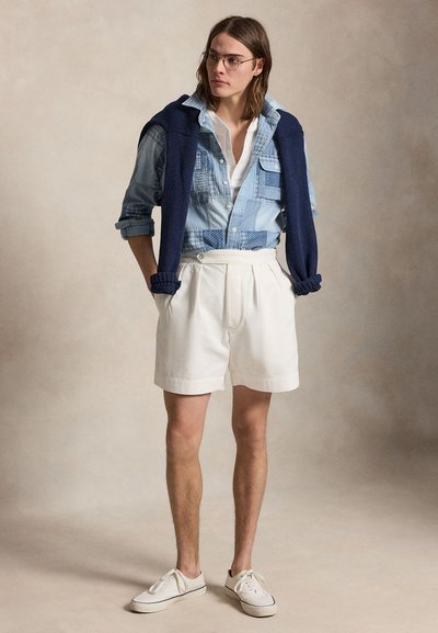 Homme aux cheveux mi-longs portant des lunettes, une chemise en jean patchwork, un short blanc, des baskets blanches et un pull bleu marine posé sur ses épaules.