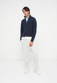 Mann står iført en marineblå quarter-zip genser, lyseblå polo, hvite bukser og hvite sneakers mot en helt hvit bakgrunn.