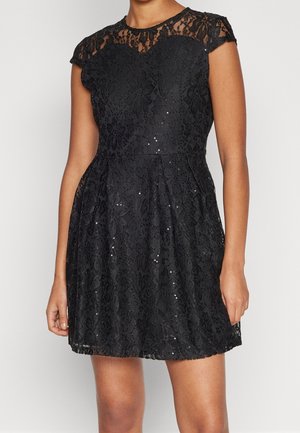 Kvinde iført en kort sort blonder kjole med kappelignende ærmer og spredte små palietter, stående mod en ensfarvet lys baggrund.