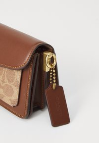 Sac à main en cuir marron avec un devant en tissu texturé beige, quincaillerie dorée et une étiquette en cuir attachée par une chaîne. Possède des coutures contrastantes.